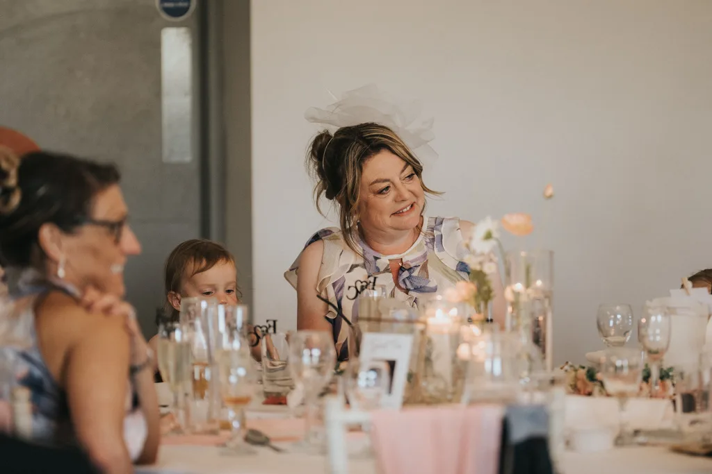 A woman in a floral dress and white fascinator smiles while seated at a candlelit, flower-adorned table at a Brackenborough wedding. Two children and another woman nearby also enjoy the formal event, captured beautifully in photography. © Aimee Lince Photography - Wedding photographer in Lincolnshire, Yorkshire & Nottinghamshire