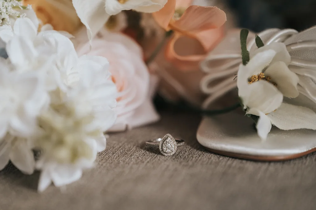 Brackenborough Lakes Resort Hotel Wedding Photos Louth Lincolnshire Chloe & Ethan 47 of 704 © Aimee Lince Photography - 03.05.2025 A teardrop-shaped diamond ring sits on a textured surface, surrounded by soft-focus white and peach flowers and a white satin shoe adorned with floral decoration and green ribbon—perfect for elegant wedding photography with a romantic, bridal atmosphere. © Aimee Lince Photography - Wedding photographer in Lincolnshire, Yorkshire & Nottinghamshire