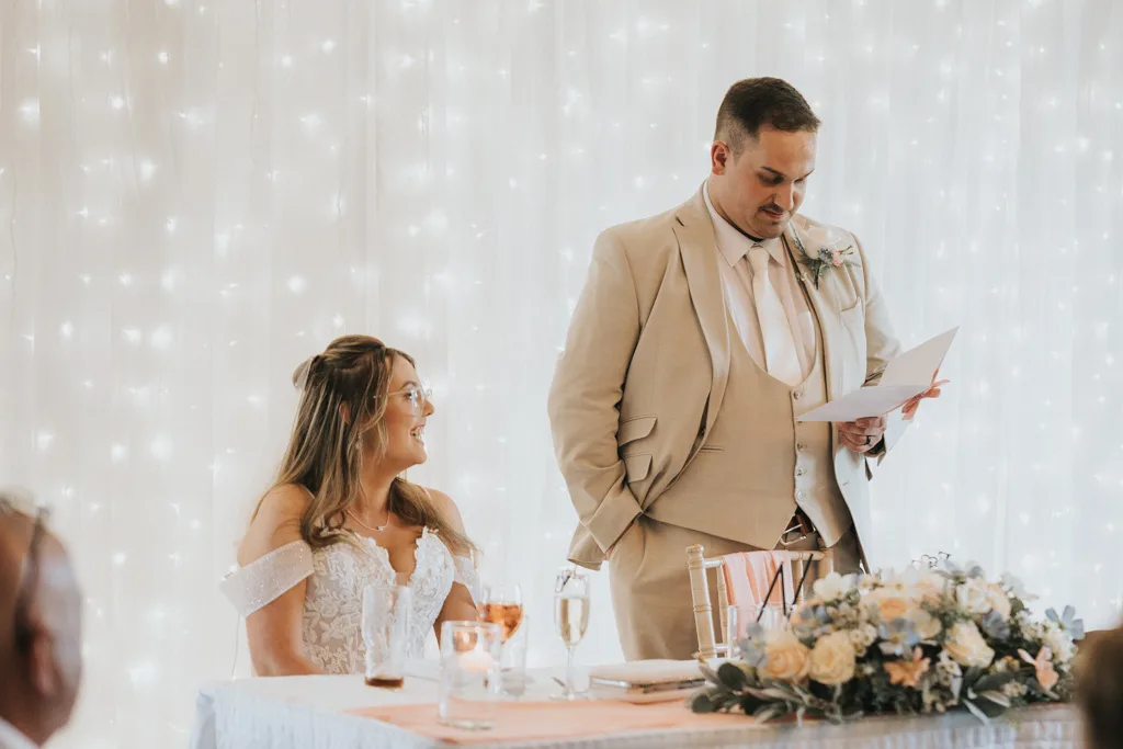 A groom in a tan suit reads from a paper beside his bride in an off-the-shoulder lace dress at their Brackenborough wedding. They sit at a table with drinks, a floral centerpiece, and soft white string lights—perfect for timeless photography. © Aimee Lince Photography - Wedding photographer in Lincolnshire, Yorkshire & Nottinghamshire