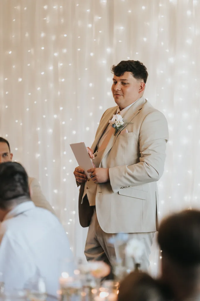 A man in a beige suit with a pale tie stands holding a piece of paper, possibly giving a wedding speech. He wears a boutonniere as out-of-focus guests and twinkling fairy lights create an elegant, festive atmosphere—perfect for Brackenborough photography. © Aimee Lince Photography - Wedding photographer in Lincolnshire, Yorkshire & Nottinghamshire