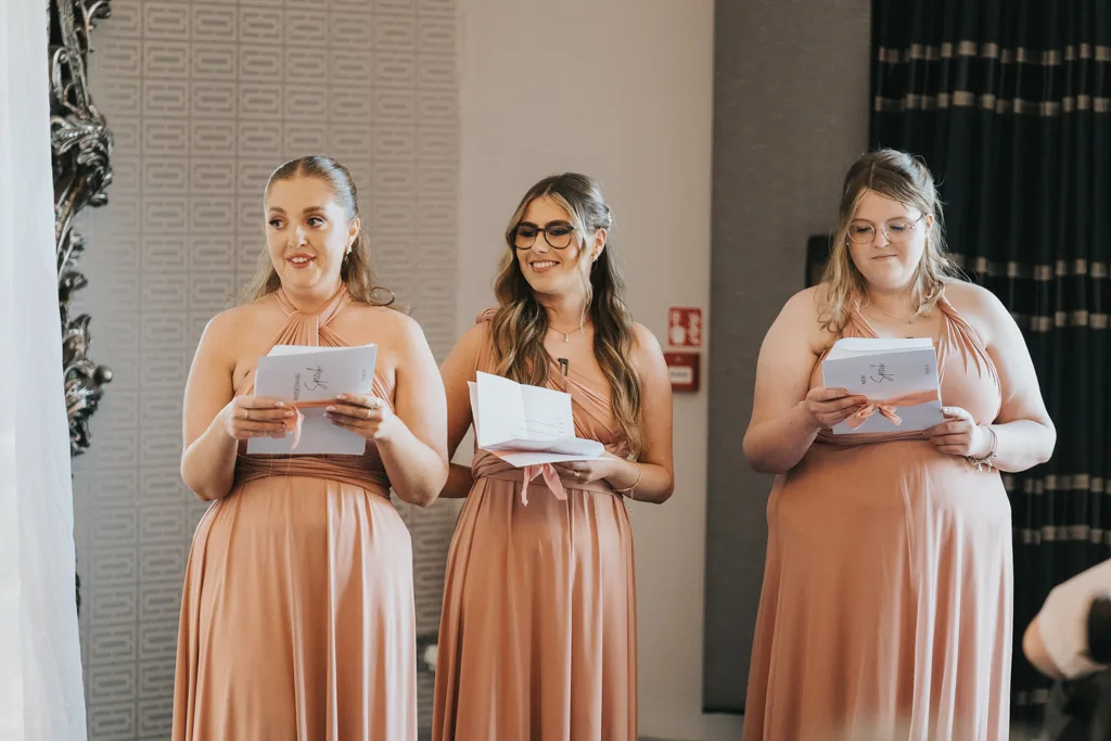 Three women in matching blush pink dresses stand indoors at Brackenborough, smiling and holding folded notes—likely bridesmaids captured during a wedding photography session. The background features patterned wallpaper, a fire extinguisher, and dark curtains. © Aimee Lince Photography - Wedding photographer in Lincolnshire, Yorkshire & Nottinghamshire
