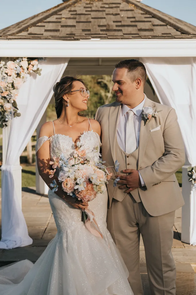 A bride and groom stand smiling at each other in front of a white gazebo decorated with flowers at Brackenborough. Sunlight highlights their happy expressions, perfectly captured in this beautiful moment of wedding photography. © Aimee Lince Photography - Wedding photographer in Lincolnshire, Yorkshire & Nottinghamshire