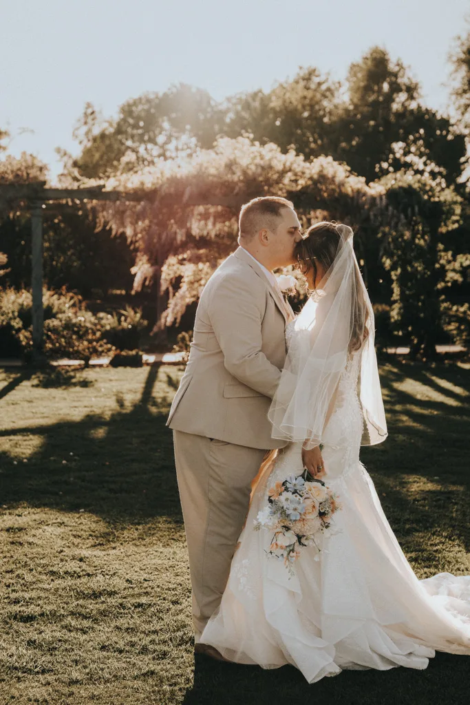 A bride in a white gown and veil holds a pastel bouquet while standing with her groom in a beige suit. Captured in stunning wedding photography, they touch foreheads in Brackenborough’s sunlit garden, surrounded by trees and flowers. © Aimee Lince Photography - Wedding photographer in Lincolnshire, Yorkshire & Nottinghamshire