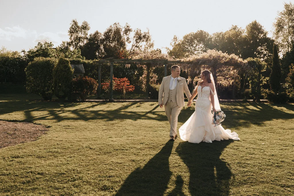 A bride and groom walk hand in hand across a sunlit lawn at Brackenborough, casting long shadows. The groom wears a light suit; the bride wears a white dress and veil, bouquet in hand—a perfect wedding photography moment under golden afternoon light. © Aimee Lince Photography - Wedding photographer in Lincolnshire, Yorkshire & Nottinghamshire