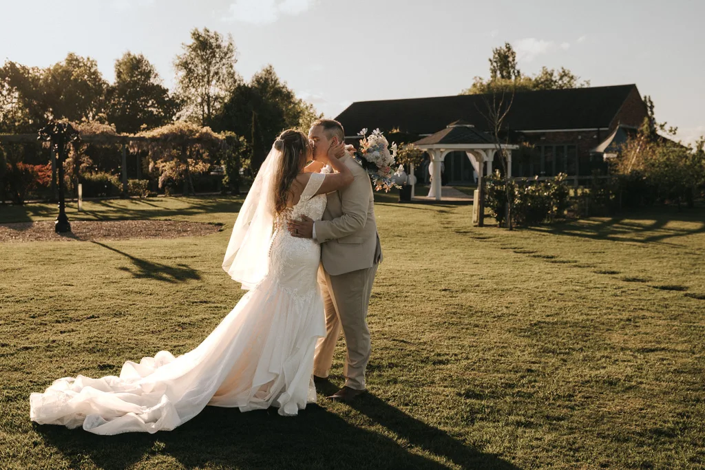 A bride in a white gown and veil and a groom in a tan suit embrace and kiss on a sunlit lawn at Brackenborough, bouquet in hand. The warm, golden light creates stunning wedding photography as trees and a gazebo cast long shadows nearby. © Aimee Lince Photography - Wedding photographer in Lincolnshire, Yorkshire & Nottinghamshire