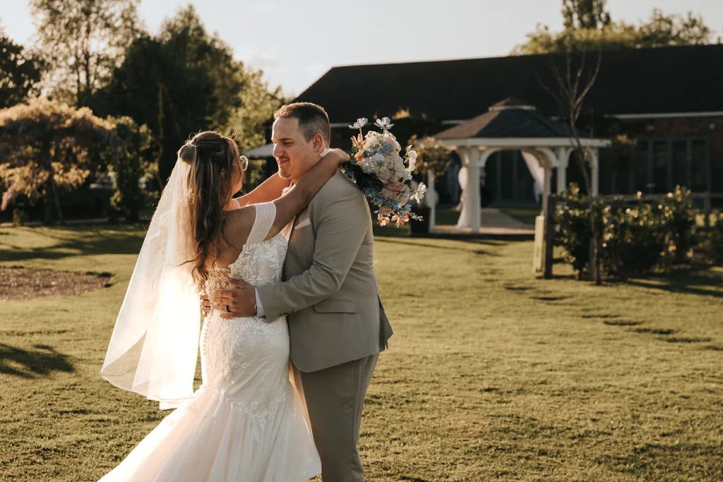 Brackenborough Lakes Resort Hotel Wedding Photos Louth Lincolnshire Chloe & Ethan 517 of 704 © Aimee Lince Photography - 03.05.2025 A bride in a white dress and veil embraces a groom in a light tan suit outdoors at Brackenborough, both smiling warmly. The bride holds a bouquet, sunlight glows behind them—a beautiful moment captured in wedding photography. © Aimee Lince Photography - Wedding photographer in Lincolnshire, Yorkshire & Nottinghamshire