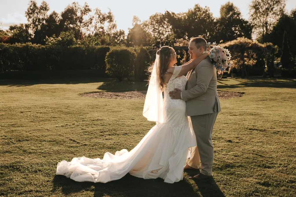 Brackenborough Lakes Resort Hotel Wedding Photos Louth Lincolnshire Chloe & Ethan 518 of 704 © Aimee Lince Photography - 03.05.2025 A bride in a long white gown and veil embraces a groom in a tan suit on the grassy Brackenborough lawn at sunset. The couple smiles at each other, bouquet in hand, as soft sunlight and trees create the perfect photography backdrop for their wedding day. © Aimee Lince Photography - Wedding photographer in Lincolnshire, Yorkshire & Nottinghamshire