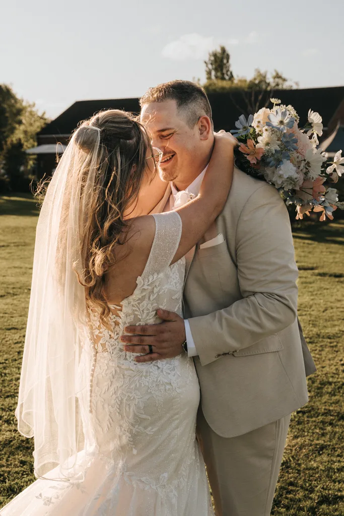 A bride and groom embrace outdoors in sunlight at Brackenborough. The bride’s white lace wedding dress and bouquet shine, while the groom dons a light beige suit. Both are smiling, forehead to forehead—captured in timeless wedding photography on lush green grass with trees behind. © Aimee Lince Photography - Wedding photographer in Lincolnshire, Yorkshire & Nottinghamshire