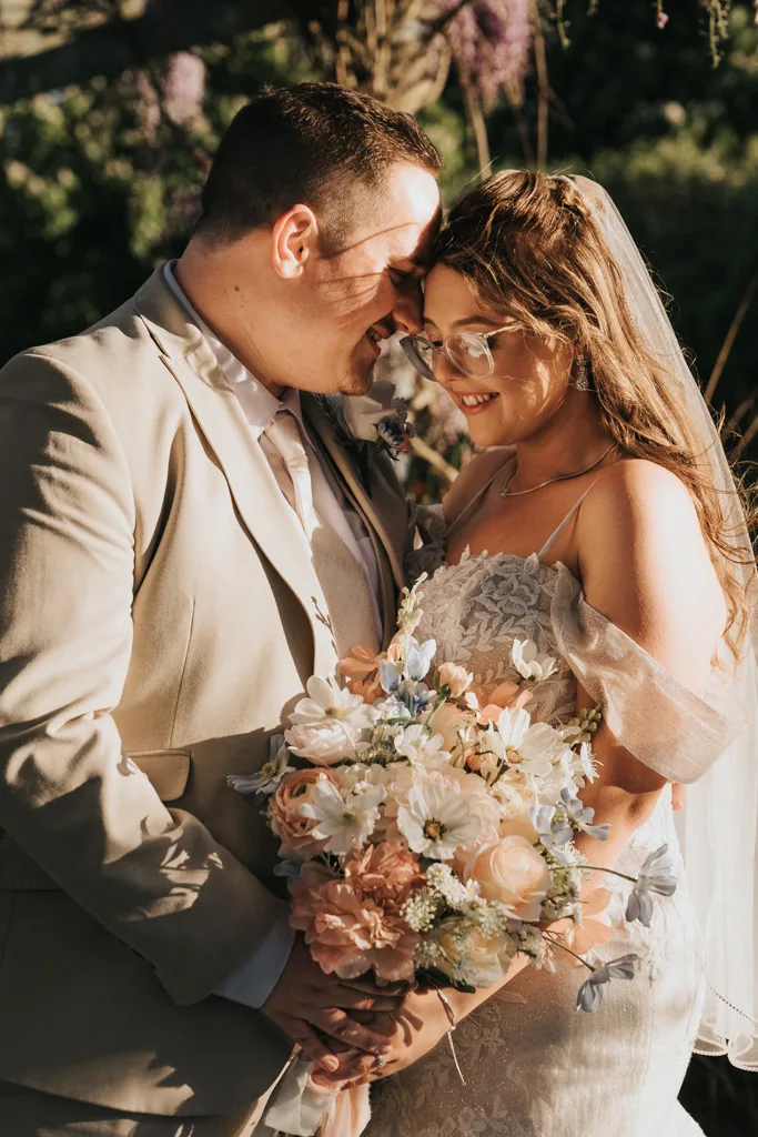 A smiling bride and groom stand close together outdoors in sunlight at Brackenborough, foreheads touching. The bride wears glasses, a lacy dress and veil, holding pastel flowers. The groom wears a light tan suit and tie. Both appear joyful and affectionate—perfect wedding photography. © Aimee Lince Photography - Wedding photographer in Lincolnshire, Yorkshire & Nottinghamshire