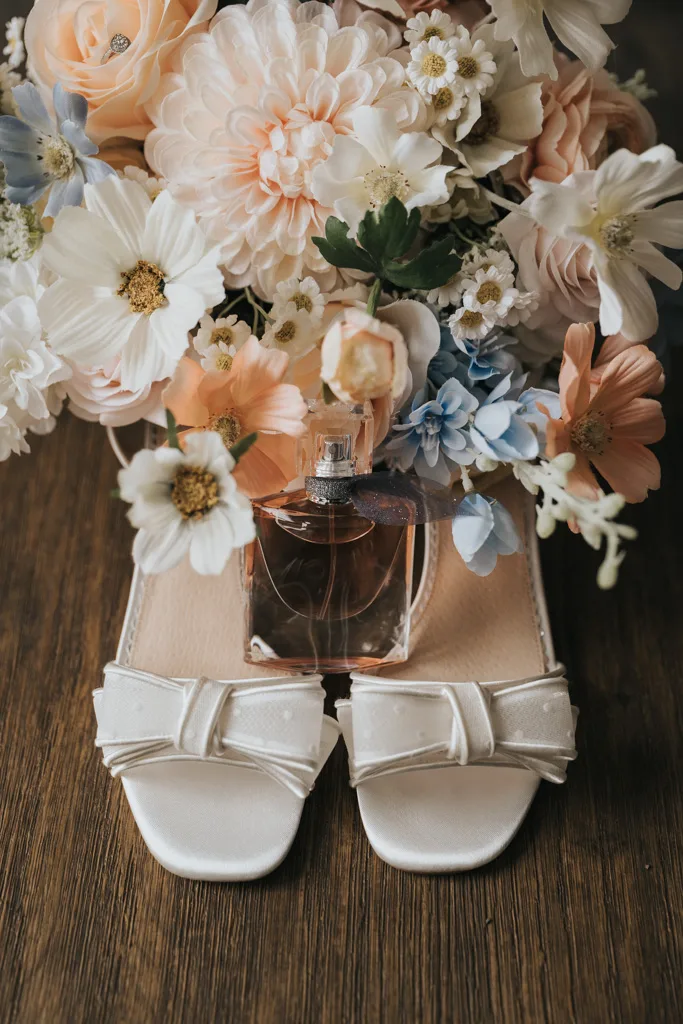 A pair of white, open-toe sandals with bow details sit on a wooden surface. A perfume bottle rests between them. Behind the shoes is a large wedding bouquet of pastel flowers, including lush foliage and soft blue blooms. © Aimee Lince Photography - Wedding photographer in Lincolnshire, Yorkshire & Nottinghamshire