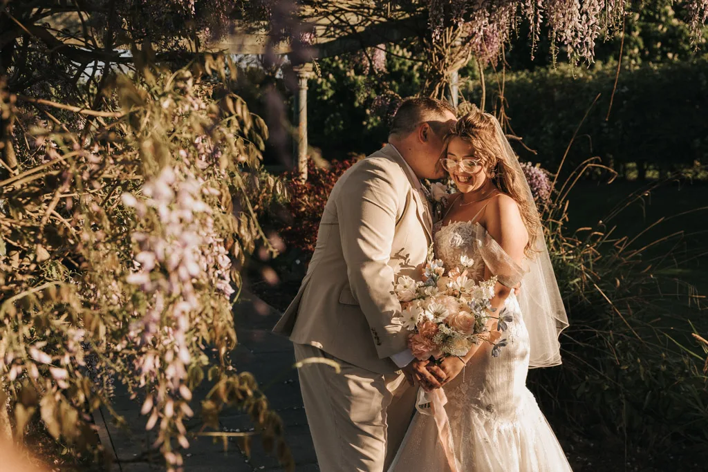 A groom in a beige suit gently kisses the smiling bride’s cheek as she holds a pastel flower bouquet. Perfect for wedding photography, they stand under a gazebo with purple wisteria at Brackenborough, surrounded by romantic natural scenery. © Aimee Lince Photography - Wedding photographer in Lincolnshire, Yorkshire & Nottinghamshire