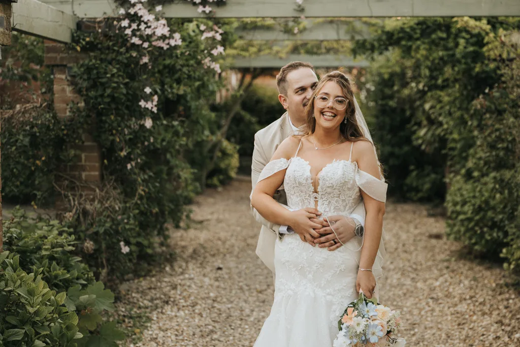 A smiling bride in a white lace dress holds a pastel bouquet while standing with her groom in a light suit. Captured by wedding photography, they embrace in a lush Brackenborough garden under wooden trellises, looking joyful together. © Aimee Lince Photography - Wedding photographer in Lincolnshire, Yorkshire & Nottinghamshire
