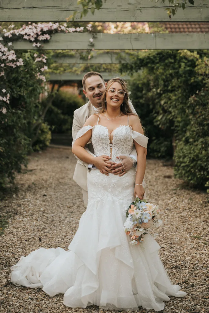 A smiling bride in a white, off-shoulder wedding dress holds a pastel bouquet, as her groom hugs her from behind amid lush greenery and flowers. This romantic garden setting at Brackenborough is perfect for unforgettable wedding photography. © Aimee Lince Photography - Wedding photographer in Lincolnshire, Yorkshire & Nottinghamshire
