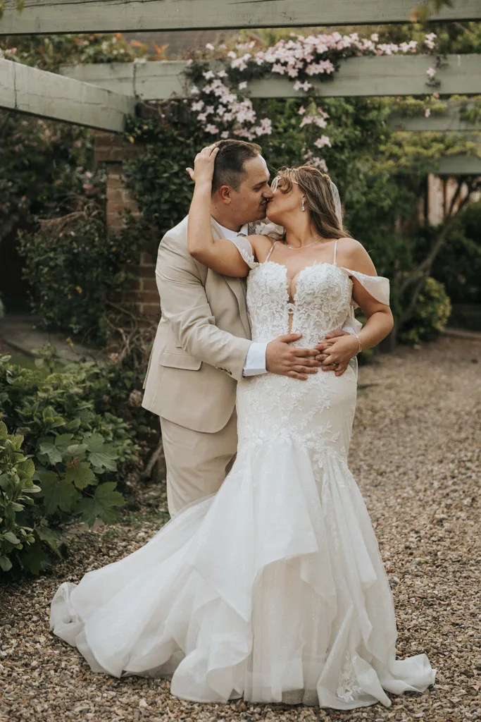 A bride and groom share a passionate kiss outdoors on their wedding day. The bride’s off-shoulder lace gown and the groom’s beige suit shine in this beautiful brackenborough photography moment under a pergola adorned with greenery and flowers. © Aimee Lince Photography - Wedding photographer in Lincolnshire, Yorkshire & Nottinghamshire