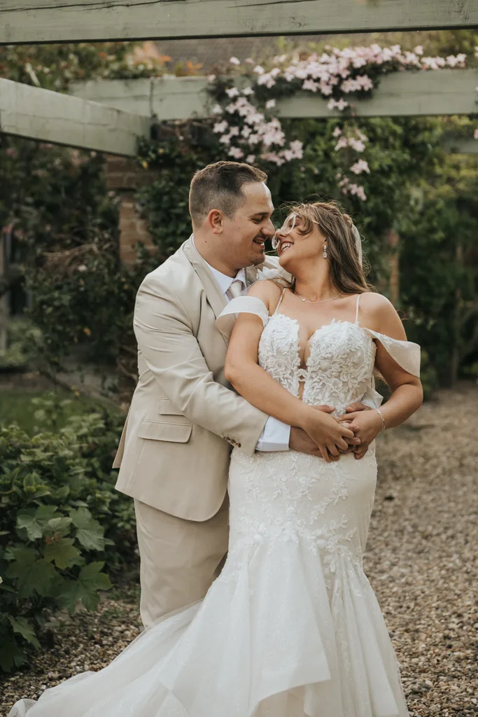 A smiling couple in wedding attire pose outdoors at Brackenborough. The man in a beige suit embraces the woman in a white lace gown. They laugh joyfully amid vibrant greenery and flowers, capturing the magic of their day through stunning wedding photography. © Aimee Lince Photography - Wedding photographer in Lincolnshire, Yorkshire & Nottinghamshire