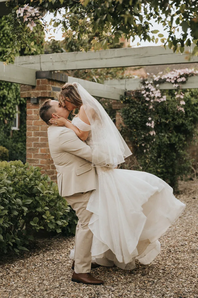 A groom in a beige suit lifts and kisses his bride in a white gown and veil under a garden pergola at Brackenborough, surrounded by greenery and brick pillars—a romantic, joyful wedding photography moment as her dress flares out beautifully. © Aimee Lince Photography - Wedding photographer in Lincolnshire, Yorkshire & Nottinghamshire