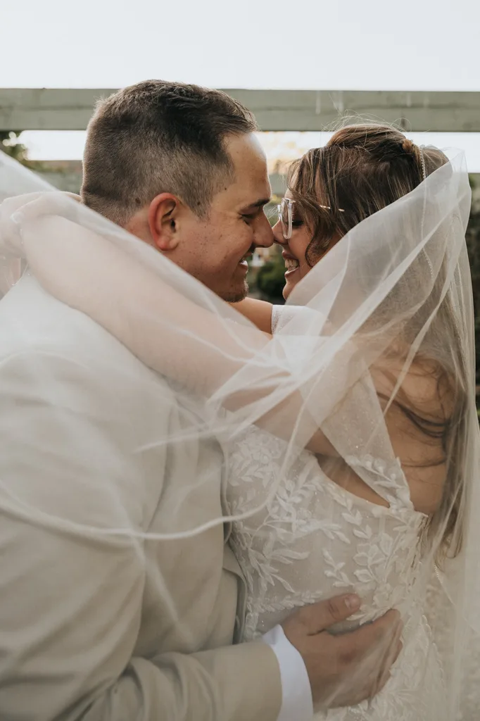 A bride and groom embrace closely, smiling at each other under the bride’s sheer veil. Captured by Brackenborough Photography, the intimate outdoor wedding moment features soft, natural lighting and a lace gown with glasses. © Aimee Lince Photography - Wedding photographer in Lincolnshire, Yorkshire & Nottinghamshire