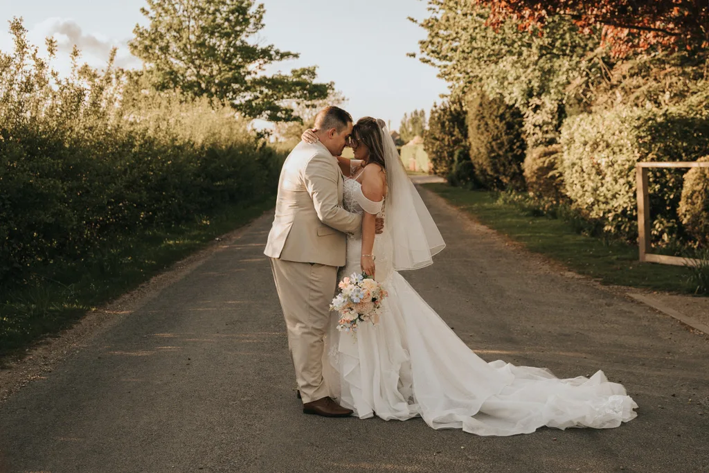 A bride and groom stand close together on a sunlit country road at Brackenborough, touching foreheads. The bride in a white gown holds a pastel bouquet, while the groom wears a beige suit—captured in romantic wedding photography amid lush greenery. © Aimee Lince Photography - Wedding photographer in Lincolnshire, Yorkshire & Nottinghamshire