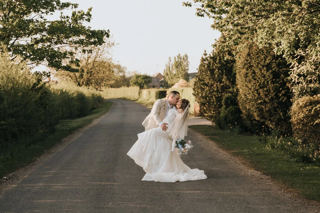 A groom in a light suit dips and kisses his bride on a quiet country road at Brackenborough, surrounded by greenery. Sunlight casts a warm glow as the couple shares a romantic wedding moment, captured beautifully in photography. © Aimee Lince Photography - Wedding photographer in Lincolnshire, Yorkshire & Nottinghamshire