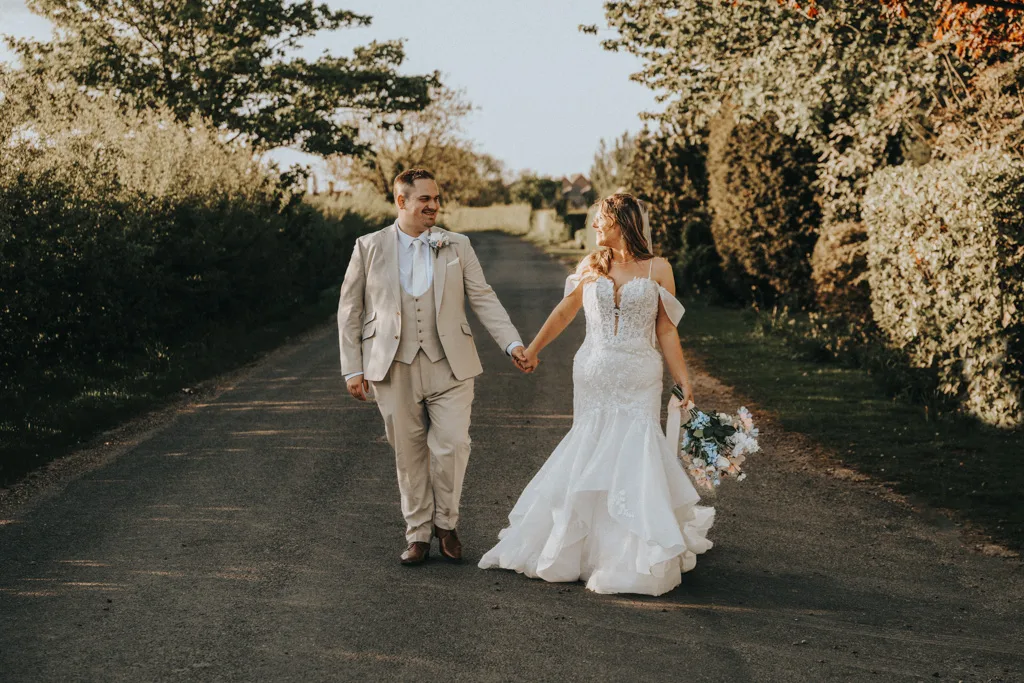 A newlywed couple walks hand in hand down a sunlit rural road at Brackenborough. The groom's light beige suit complements the bride’s elegant gown and bouquet, as lush greenery frames this charming wedding photography moment. © Aimee Lince Photography - Wedding photographer in Lincolnshire, Yorkshire & Nottinghamshire