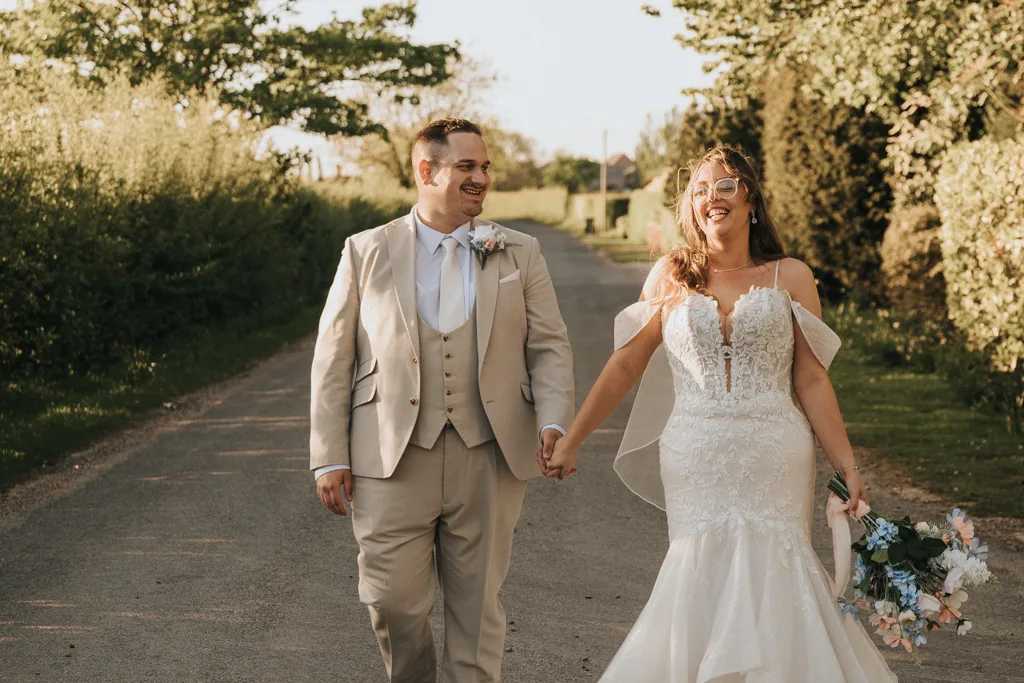 A couple in wedding attire walk hand in hand down a sunlit country road at Brackenborough. The groom wears a beige suit with a white shirt and tie; the bride, in a fitted lace gown, smiles and holds a bouquet—perfect for timeless wedding photography. © Aimee Lince Photography - Wedding photographer in Lincolnshire, Yorkshire & Nottinghamshire