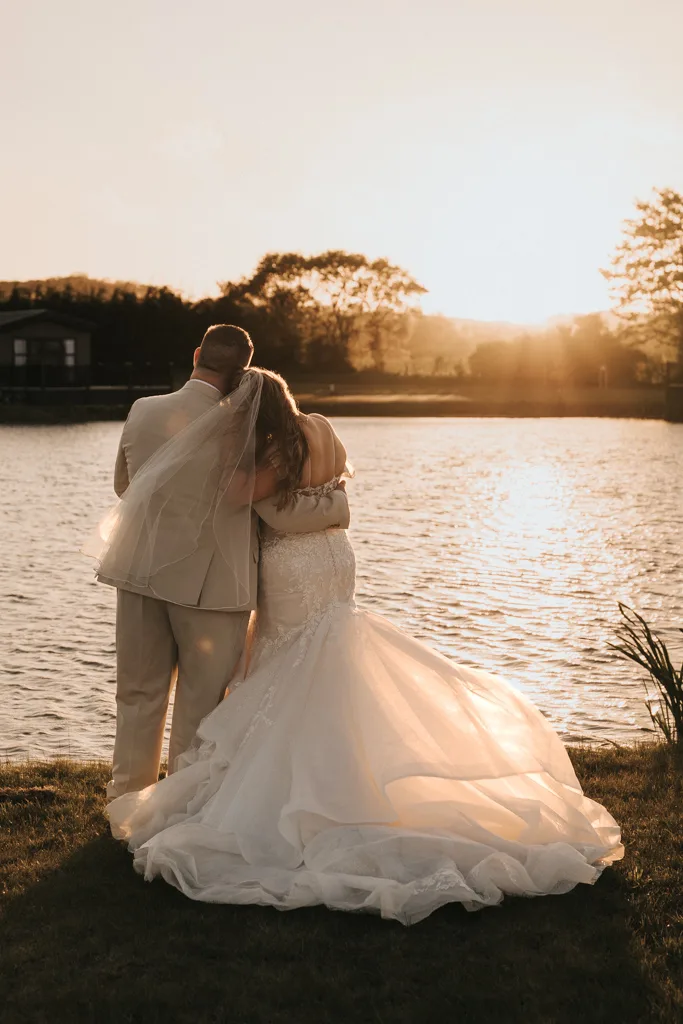 A bride and groom stand by a lakeshore at sunset, embracing and facing the water. The wedding photography captures warm sunlight glowing around them, with reflections on the calm lake at Brackenborough. © Aimee Lince Photography - Wedding photographer in Lincolnshire, Yorkshire & Nottinghamshire