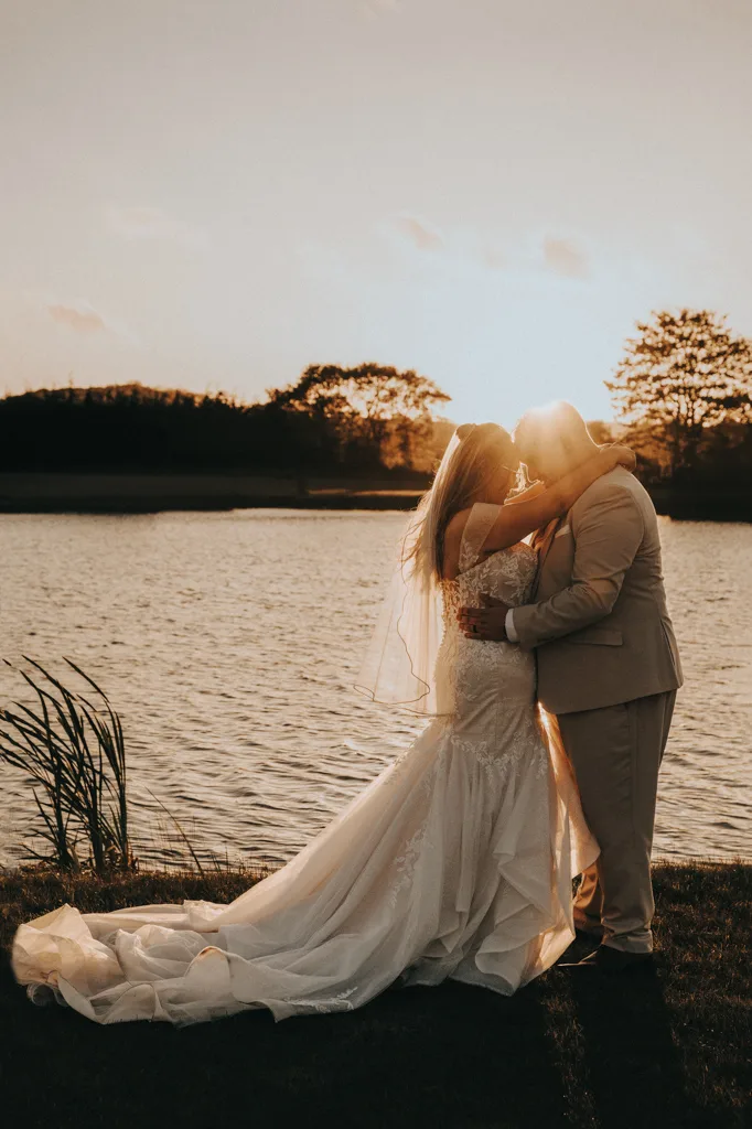 A bride and groom embrace by a lake at sunset, their wedding attire glowing in the golden light. Captured through enchanting photography, the romantic scene at Brackenborough radiates warmth as they lovingly touch foreheads. © Aimee Lince Photography - Wedding photographer in Lincolnshire, Yorkshire & Nottinghamshire