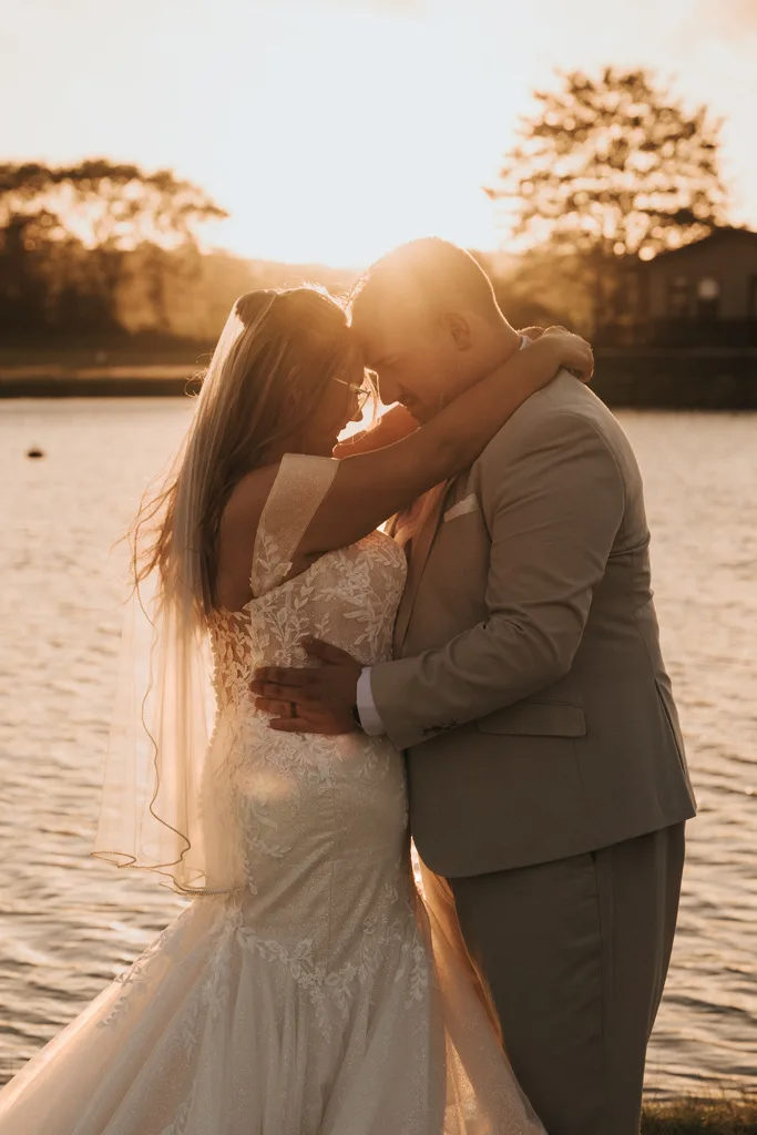 A bride and groom embrace by a lakeside at sunset in this stunning wedding photography at Brackenborough; warm sunlight creates a soft glow as their foreheads touch, with rippling water and trees framing the romantic moment. © Aimee Lince Photography - Wedding photographer in Lincolnshire, Yorkshire & Nottinghamshire