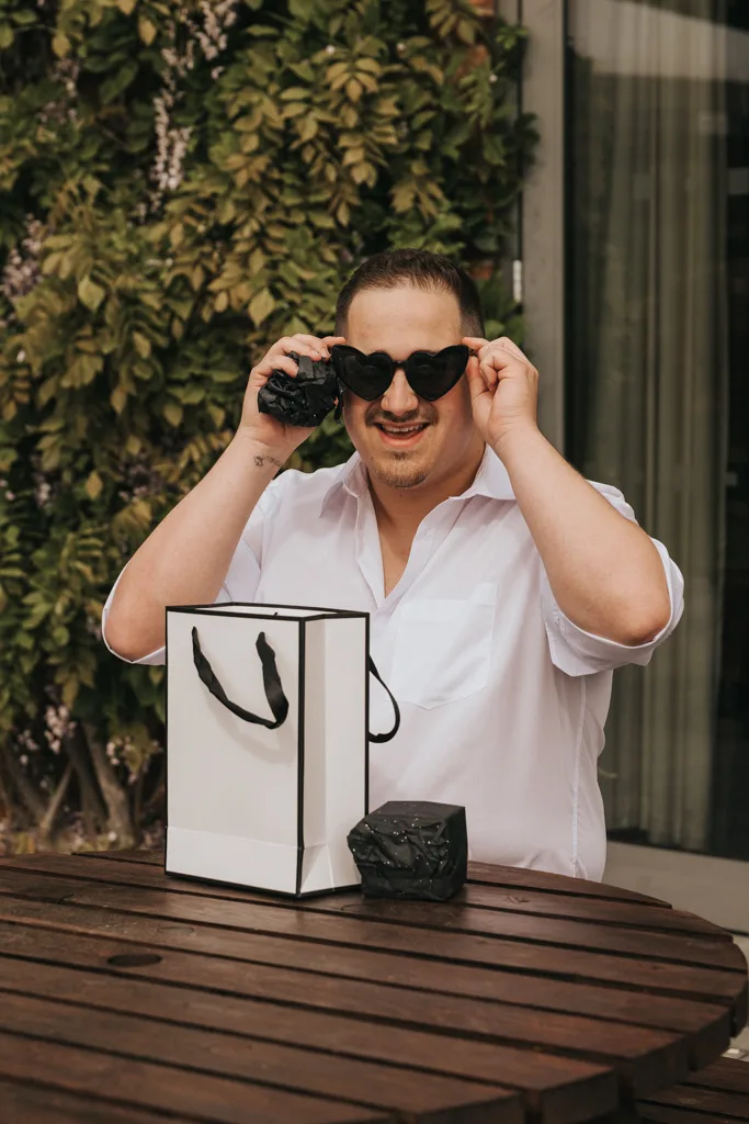 A smiling man in a white shirt sits at a wooden table outdoors at Brackenborough, playfully holding his heart-shaped sunglasses. In front of him are a white gift bag and a black-wrapped box—perfect for candid wedding photography amid lush greenery. © Aimee Lince Photography - Wedding photographer in Lincolnshire, Yorkshire & Nottinghamshire