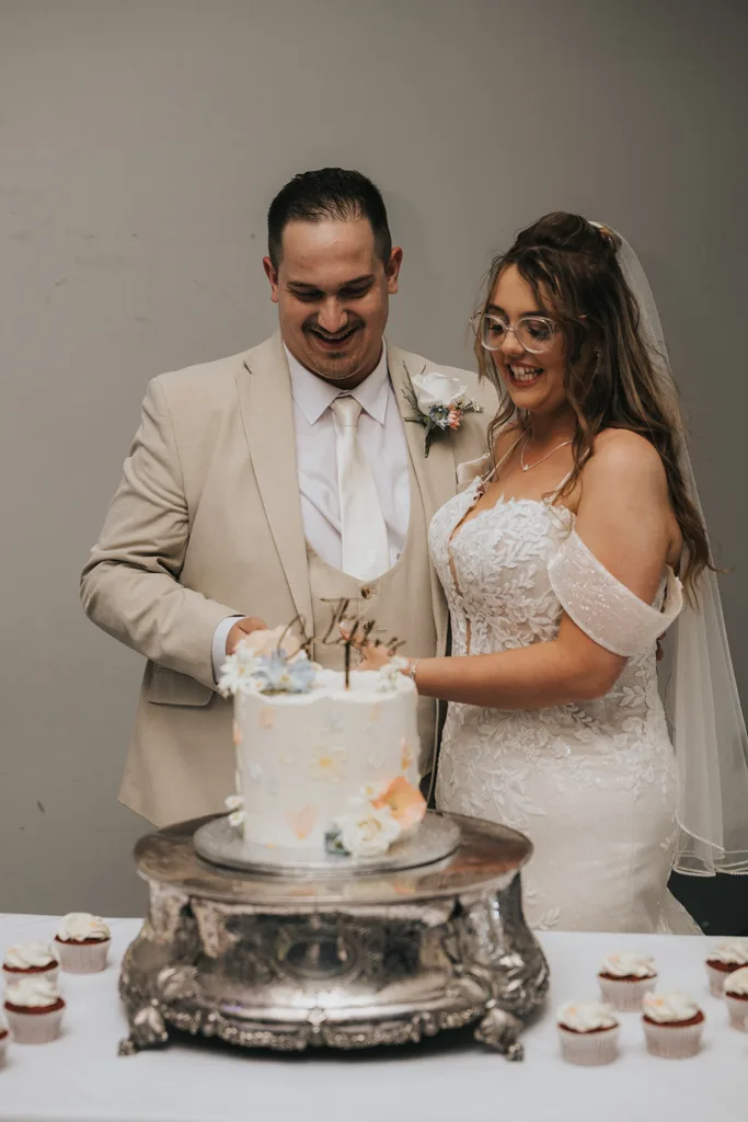 At their Brackenborough wedding, a bride in an off-shoulder lace gown and veil stands with her groom by a white tiered cake. Smiling for the photography, they cut the floral cake surrounded by cupcakes on a white tablecloth. © Aimee Lince Photography - Wedding photographer in Lincolnshire, Yorkshire & Nottinghamshire