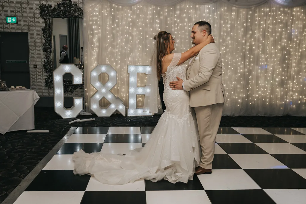 At a Brackenborough wedding, the bride and groom dance on a black-and-white checkered floor. The illuminated "C&E" letters and fairy lights create a magical scene, perfectly captured in this stunning photography. © Aimee Lince Photography - Wedding photographer in Lincolnshire, Yorkshire & Nottinghamshire