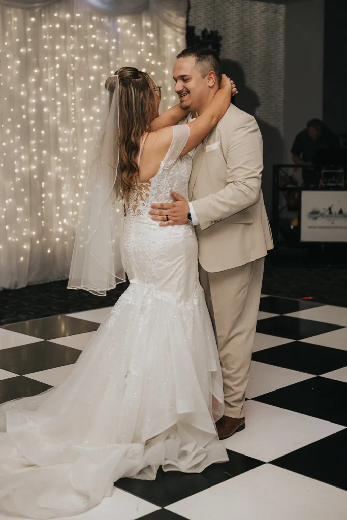 A bride in a white lace gown and veil dances with her groom in a tan suit on a black-and-white checkered floor at their Brackenborough wedding, captured through stunning photography beneath sparkling fairy lights for a truly romantic ambiance. © Aimee Lince Photography - Wedding photographer in Lincolnshire, Yorkshire & Nottinghamshire