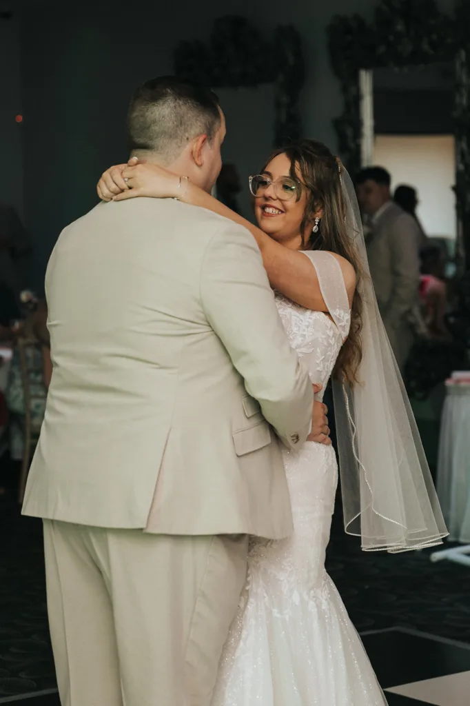 A bride in a white lace wedding dress and veil smiles warmly at her groom during their first dance. The groom, in a light beige suit, holds her gently. Brackenborough wedding photography captures guests and tables softly lit in the blurred background. © Aimee Lince Photography - Wedding photographer in Lincolnshire, Yorkshire & Nottinghamshire