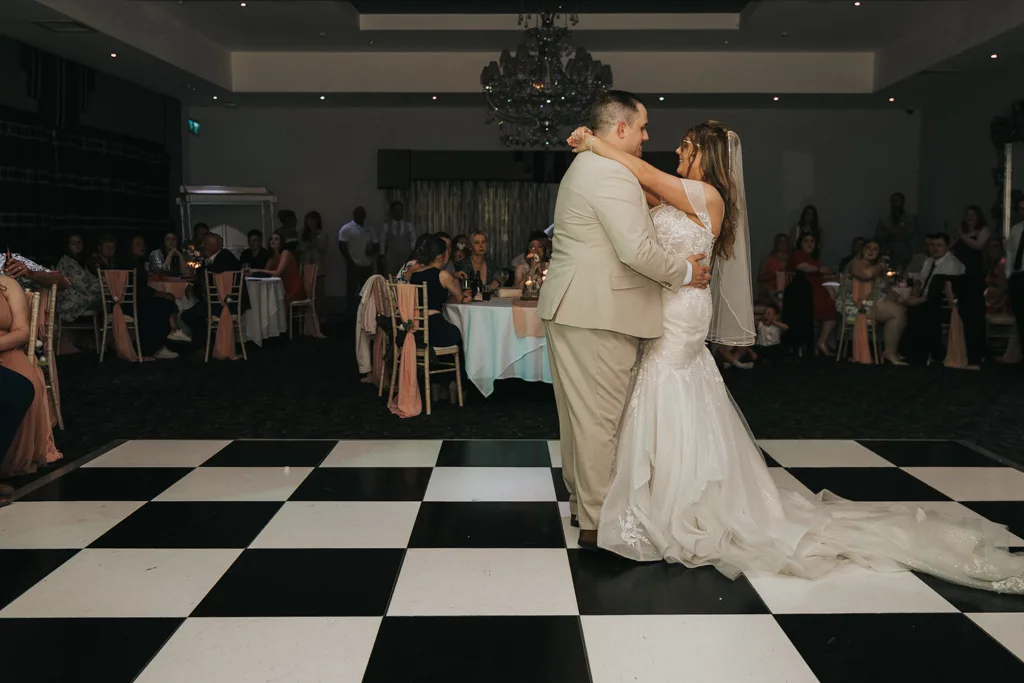 At Brackenborough, wedding photography captures a bride and groom’s first dance on a checkered floor, surrounded by guests in an elegant, dimly lit reception hall. The bride’s long train and groom’s light suit complete the romantic scene. © Aimee Lince Photography - Wedding photographer in Lincolnshire, Yorkshire & Nottinghamshire