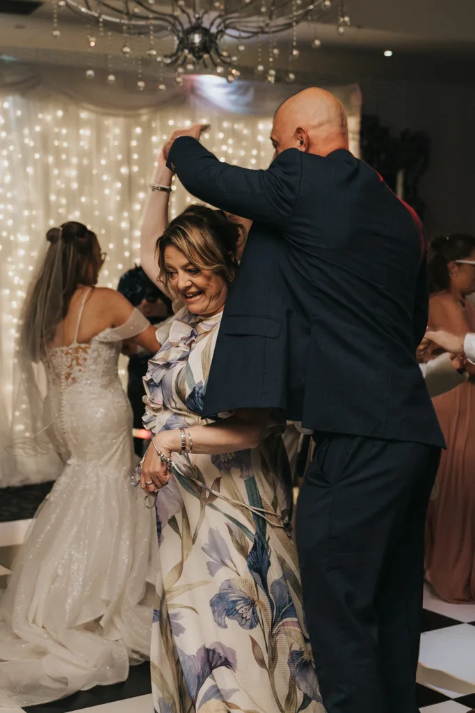 A man in a dark suit twirls a smiling woman in a floral dress on the dance floor at a wedding. Behind them, the bride dances beneath soft, warm lights and draped fabric, creating an elegant photography scene at Brackenborough. © Aimee Lince Photography - Wedding photographer in Lincolnshire, Yorkshire & Nottinghamshire
