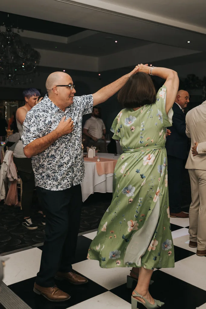 A man in a floral shirt and glasses dances with a woman in a green, floral dress and heels on the black-and-white checkered floor at a Brackenborough wedding, while others mingle near tables draped in white tablecloths—perfect for photography. © Aimee Lince Photography - Wedding photographer in Lincolnshire, Yorkshire & Nottinghamshire