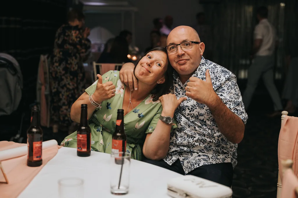 A smiling woman in a green floral dress and a bald man in glasses and a patterned shirt sit close together at a table, both giving thumbs up. Wedding photography captures beer bottles, napkins, and other guests at Brackenborough in the dimly lit room. © Aimee Lince Photography - Wedding photographer in Lincolnshire, Yorkshire & Nottinghamshire
