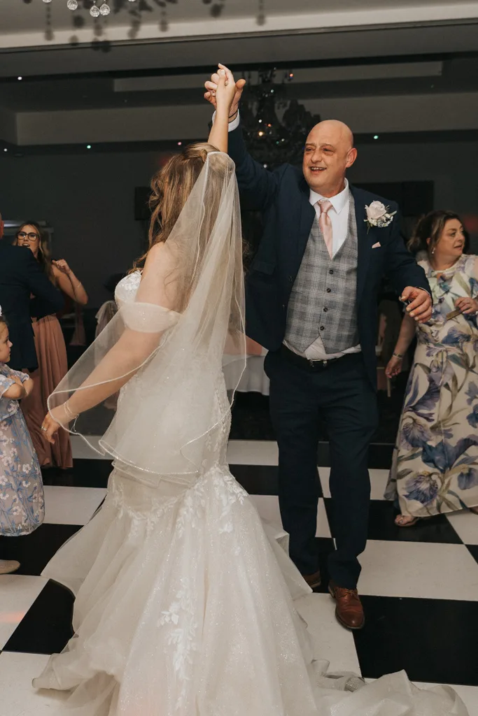 A bride in a white gown and veil dances with a smiling, bald man in a navy suit and pink tie on a black-and-white checkered floor. Guests watch and enjoy the Brackenborough wedding reception, beautifully captured in this vibrant photography. © Aimee Lince Photography - Wedding photographer in Lincolnshire, Yorkshire & Nottinghamshire