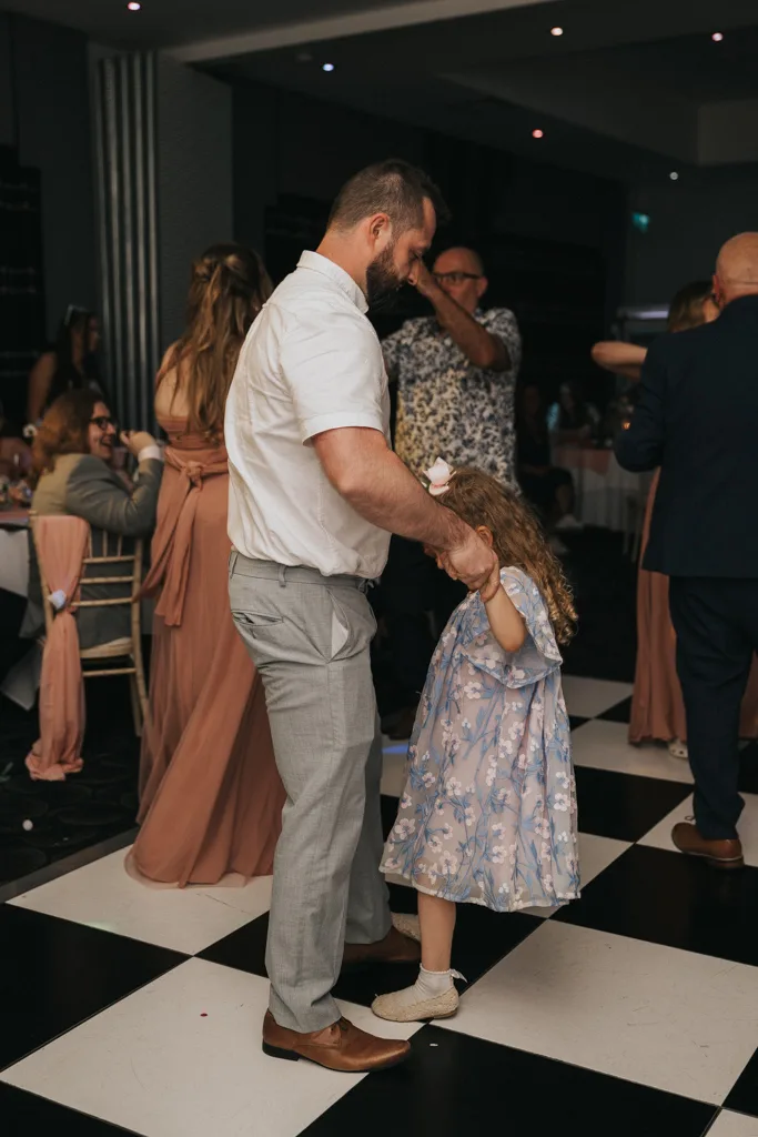 A man in a white shirt and gray pants dances hand-in-hand with a young girl in a blue floral dress on the checkered floor at a lively wedding reception. Guests in formal attire surround them, capturing joyful moments through photography. © Aimee Lince Photography - Wedding photographer in Lincolnshire, Yorkshire & Nottinghamshire