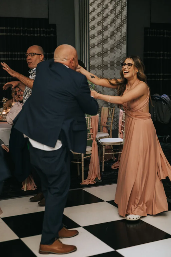 A smiling woman in a peach gown and white Crocs dances with a bald man in a blue suit on a checkered floor at a Brackenborough wedding. Nearby, another man and woman watch as the moment is captured in vibrant photography amid elegant, dark decor. © Aimee Lince Photography - Wedding photographer in Lincolnshire, Yorkshire & Nottinghamshire