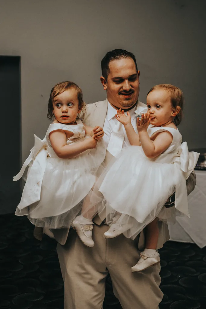 A man in a tan suit smiles while holding two young twin girls in white dresses and matching shoes, one in each arm. Captured at a wedding, this brackenborough photography moment features the girls with light brown hair looking in different directions. © Aimee Lince Photography - Wedding photographer in Lincolnshire, Yorkshire & Nottinghamshire