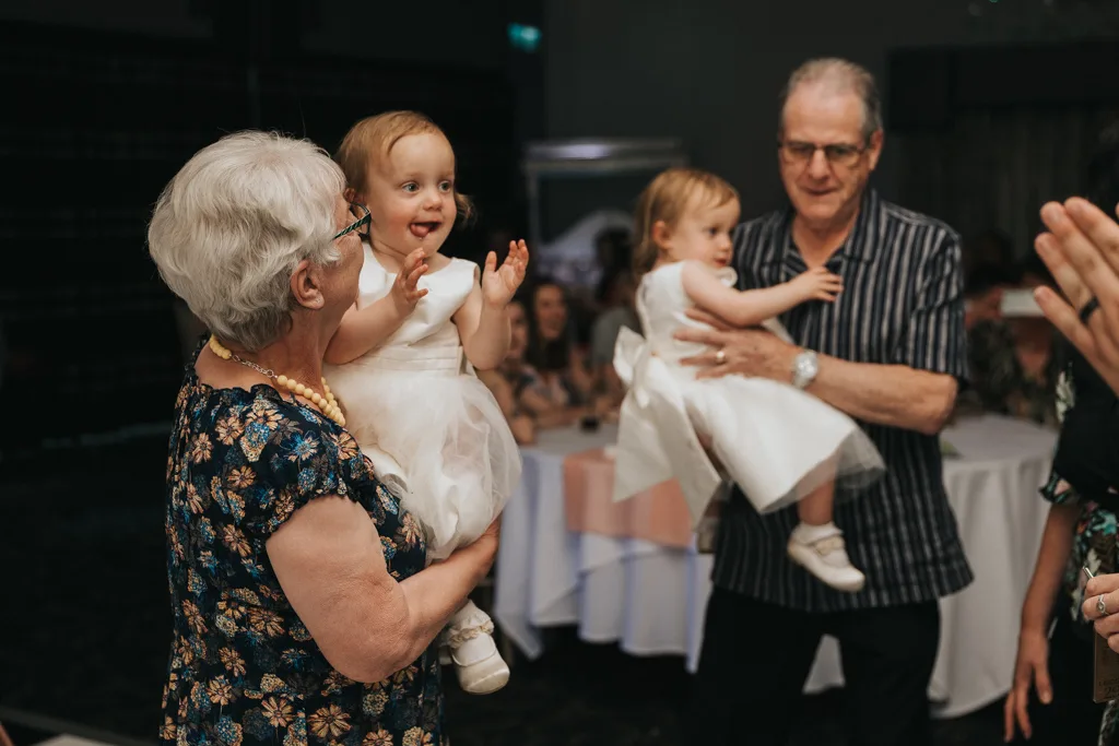 Two smiling older adults each hold a happy toddler girl in a white dress and shoes. Indoors at a Brackenborough wedding celebration, the group poses for photography as pink decorations and seated guests are visible in the background. © Aimee Lince Photography - Wedding photographer in Lincolnshire, Yorkshire & Nottinghamshire
