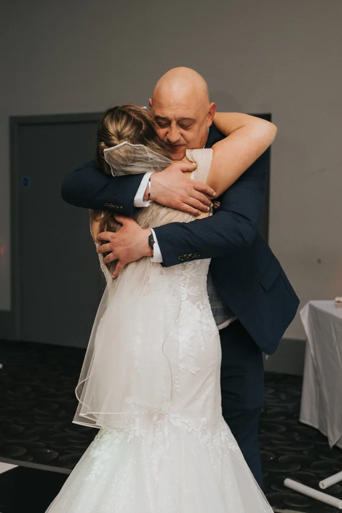 A bride in a white gown and veil hugs an older, bald man in a suit at a wedding. They embrace tightly, eyes closed, conveying deep emotion. The setting appears to be indoors with simple décor—a beautiful moment of wedding photography. © Aimee Lince Photography - Wedding photographer in Lincolnshire, Yorkshire & Nottinghamshire