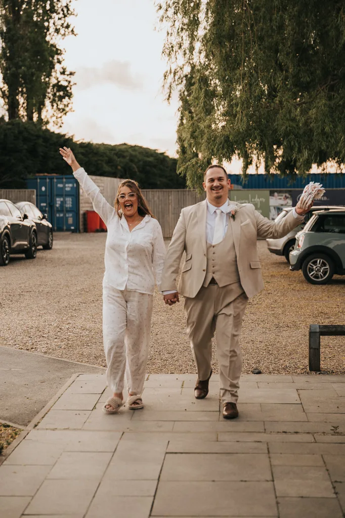A joyful couple walks hand in hand outdoors, smiling widely. The woman wears white pajamas and slippers, the man a beige suit and tie—captured in candid wedding photography. Parked cars and large blue shipping containers are in the background. © Aimee Lince Photography - Wedding photographer in Lincolnshire, Yorkshire & Nottinghamshire