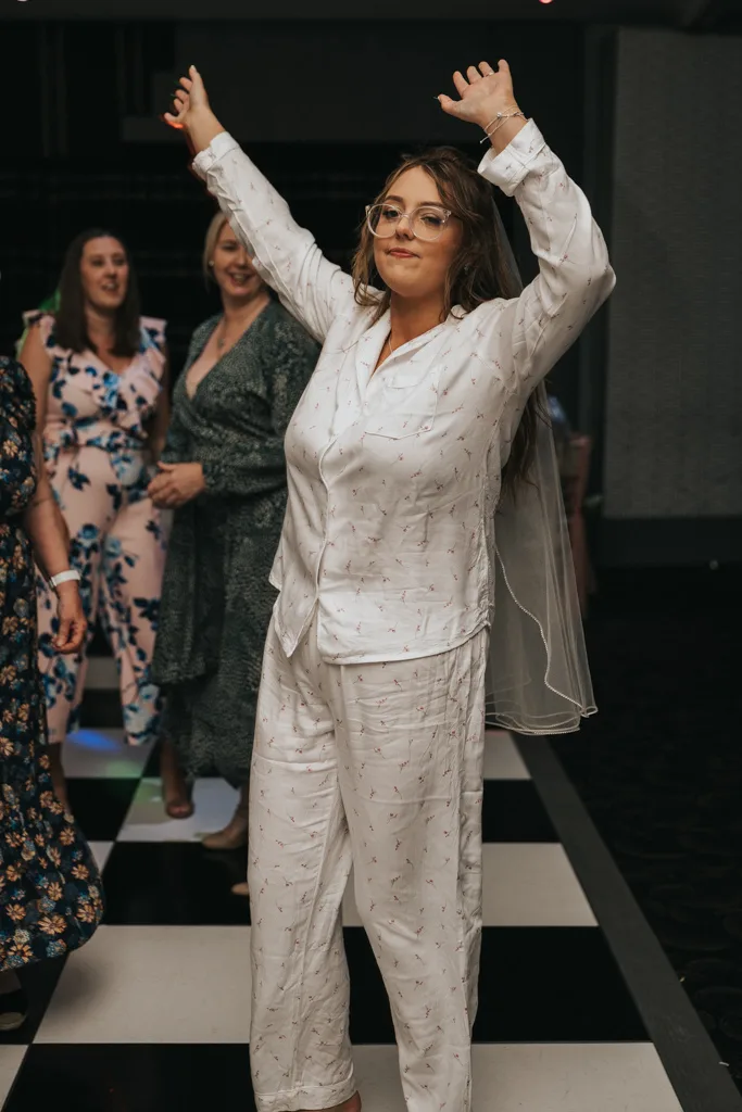A woman in white pajamas, glasses, and a bridal veil stands on a black-and-white checkered floor at what appears to be a Brackenborough wedding. She smiles with arms raised as three women in patterned dresses watch, capturing a joyful moment in photography. © Aimee Lince Photography - Wedding photographer in Lincolnshire, Yorkshire & Nottinghamshire