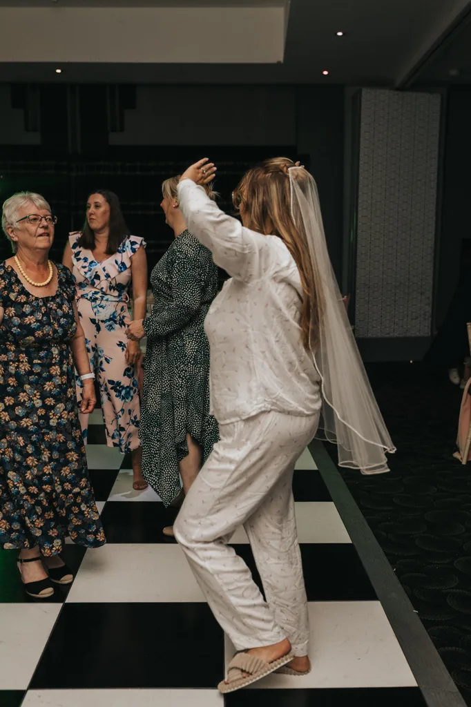 A woman in white pajamas and a bridal veil dances on a black-and-white checkered floor during a joyful wedding celebration at Brackenborough. Three other women in floral dresses stand nearby, watching her. The photography captures the festive indoor scene perfectly. © Aimee Lince Photography - Wedding photographer in Lincolnshire, Yorkshire & Nottinghamshire