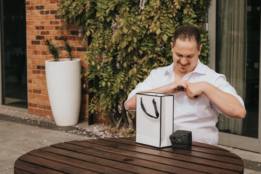 Brackenborough Lakes Resort Hotel Wedding Photos Louth Lincolnshire Chloe & Ethan 8 of 704 © Aimee Lince Photography - 03.05.2025 A man wearing a white shirt sits at a round wooden table outdoors, smiling as he opens a white gift bag with black handles during wedding photography at Brackenborough. Behind him are a brick wall, green vines, and a large white planter. © Aimee Lince Photography - Wedding photographer in Lincolnshire, Yorkshire & Nottinghamshire