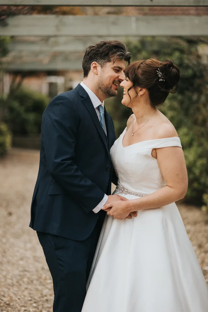 A bride in an off-shoulder white gown holds hands and smiles closely with a groom in a navy suit and tie outdoors, under a wooden pergola with greenery in the background, both appearing joyful and affectionate. © Aimee Lince Photography - Wedding photographer in Lincolnshire, Yorkshire & Nottinghamshire