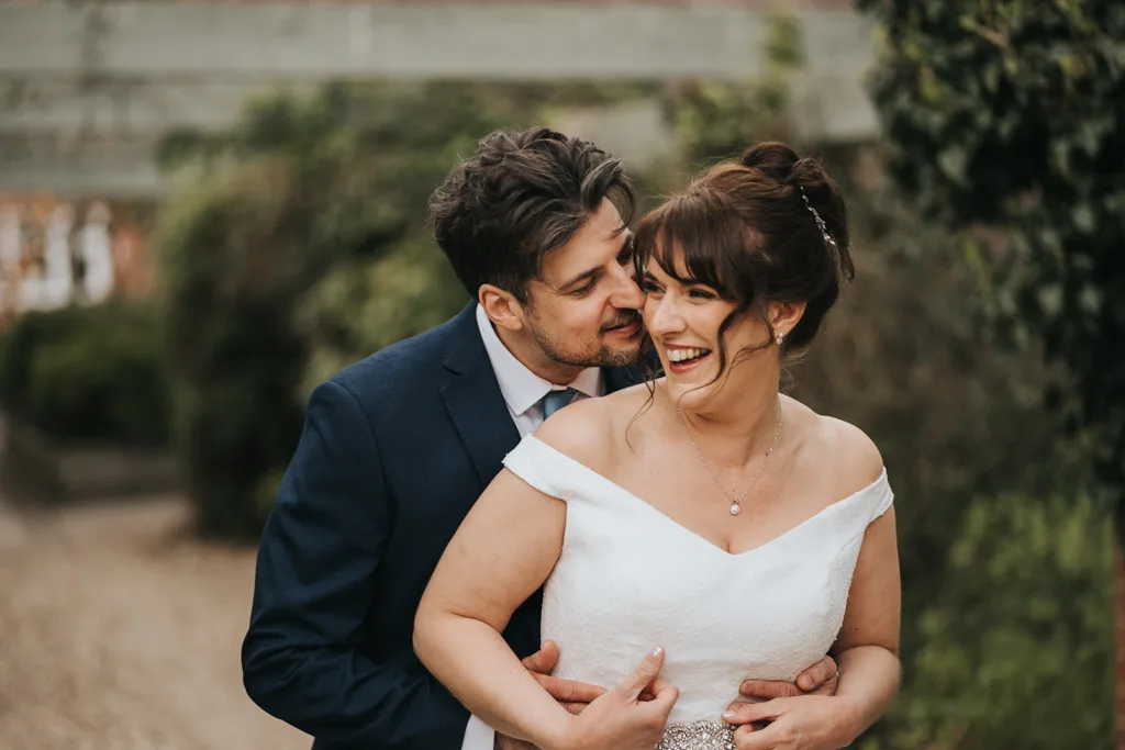 A groom in a navy suit embraces a smiling bride in an off-shoulder white dress from behind. Both are laughing joyfully outdoors, surrounded by greenery, with soft natural light and blurred foliage in the background. © Aimee Lince Photography - Wedding photographer in Lincolnshire, Yorkshire & Nottinghamshire