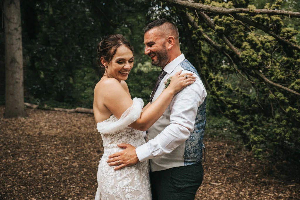 A bride in a strapless, lace wedding dress and a groom in a white shirt and patterned vest embrace and laugh together outdoors, surrounded by trees and greenery, with natural light highlighting their joyful expressions. © Aimee Lince Photography - Wedding photographer in Lincolnshire, Yorkshire & Nottinghamshire